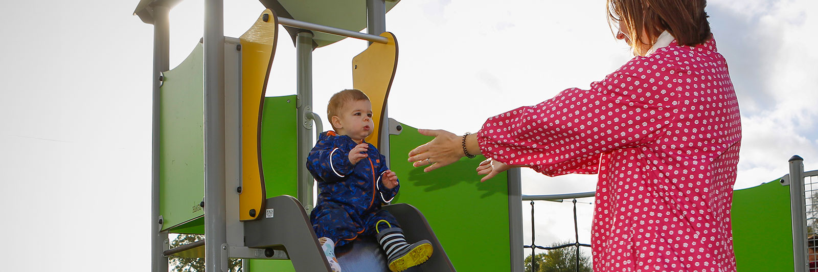 An adult holds her arms out for a toddler standing on top of a playground unit at the top of the slide.